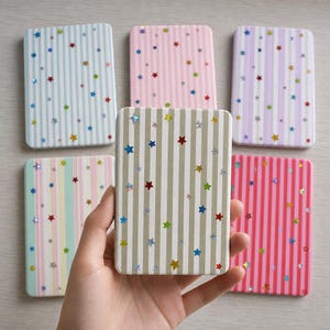 Könnte beinhalten: Fünf rechteckige Taschenspiegel mit vertikalen Streifen in verschiedenen Pastellfarben wie Hellblau, Rosa, Lavendel und Beige. Jeder Spiegel ist mit kleinen, verstreuten holografischen Sternen auf der gesamten Oberfläche verziert.