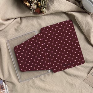 May include: A dark maroon tablet case featuring a repeating pattern of small light pink hearts. The case has a folding cover design and includes the name Brandon printed in a cursive script near the bottom right corner of the front panel.