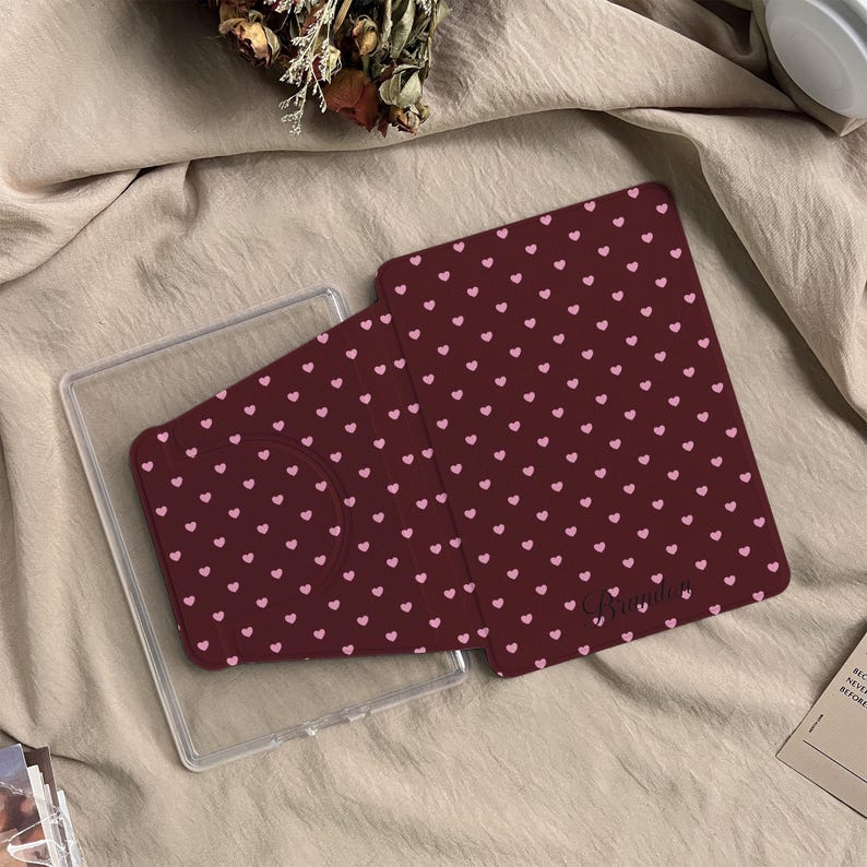 May include: A dark maroon tablet case featuring a repeating pattern of small light pink hearts. The case has a folding cover design and includes the name Brandon printed in a cursive script near the bottom right corner of the front panel.
