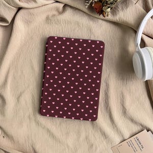 May include: A rectangular e-reader case featuring a dark maroon background with a repeating pattern of small, light pink hearts. The case is positioned on a beige fabric surface next to white over-ear headphones and a small card with text.