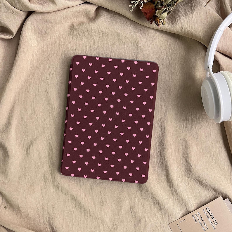 May include: A rectangular e-reader case featuring a dark maroon background with a repeating pattern of small, light pink hearts. The case is positioned on a beige fabric surface next to white over-ear headphones and a small card with text.