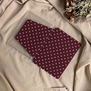 May include: A dark maroon e-reader or tablet case featuring a repeating pattern of small light pink hearts. The case includes a clear plastic frame component and a folding cover with a textured surface, designed to protect the device screen.