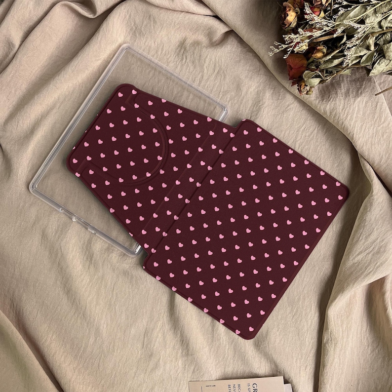 May include: A dark maroon e-reader or tablet case featuring a repeating pattern of small light pink hearts. The case includes a clear plastic frame component and a folding cover with a textured surface, designed to protect the device screen.