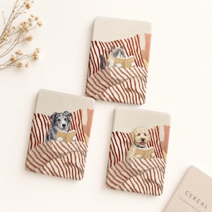 Puede incluir: Tres fundas rectangulares para lector electrónico con ilustraciones de un gato, un perro gris y un perro dorado, cada uno en la cama con mantas de rayas rojas y blancas mientras leen libros. Se ve la esquina de un libro titulado CEREAL.