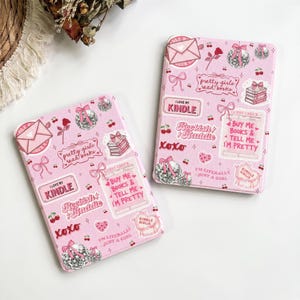 Puede incluir: Dos fundas para lector electrónico de color rosa claro con un patrón repetitivo de estilo pegatina de bolas de discoteca, lazos, cerezas, rosas, sobres y pilas de libros. El texto incluye "I love my Kindle" y "Buy me books".