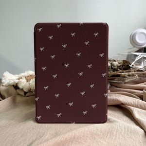 Puede incluir: Una funda rectangular para lector electr&oacute;nico en color burdeos intenso con un patr&oacute;n repetitivo de peque&ntilde;os lazos finos dibujados en blanco, dispersos por toda la superficie. La funda tiene un acabado mate liso y esquinas redondeadas.