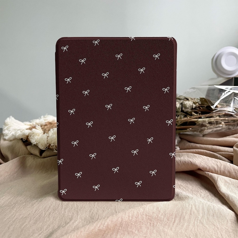 Puede incluir: Una funda rectangular para lector electr&oacute;nico en color burdeos intenso con un patr&oacute;n repetitivo de peque&ntilde;os lazos finos dibujados en blanco, dispersos por toda la superficie. La funda tiene un acabado mate liso y esquinas redondeadas.