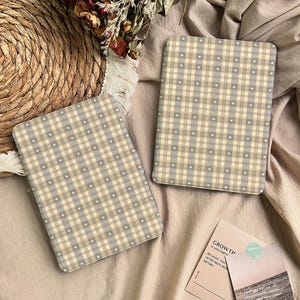 Può includere: Due custodie rettangolari con un motivo a quadri beige, grigio e bianco, decorate con piccoli cuori bianchi. Le custodie sono esposte su una superficie di tessuto beige, con un cestino intrecciato e fiori secchi sullo sfondo. È visibile anche una cartolina con la parola "GROWTH".