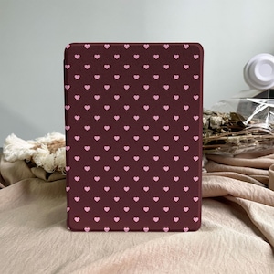 May include: A rectangular e-reader case featuring a dark maroon background with a repeating pattern of small, light pink hearts arranged in uniform rows and columns across the entire front surface of the protective cover.