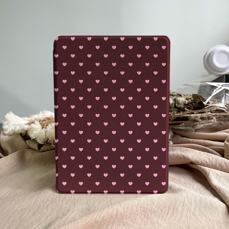 May include: A rectangular e-reader case featuring a dark maroon background with a repeating pattern of small, light pink hearts arranged in uniform rows and columns across the entire front surface of the protective cover.