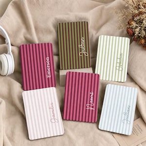 May include: Six rectangular notebooks with vertical striped covers in various colours including maroon, olive green, light green, light pink, and light blue. Each notebook features a white script name printed on the front: Emerson, Julian, Ophelia, Jamilia, Naomi, and Julian.