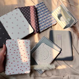 Può includere: Una collezione di custodie per tablet con diversi motivi. I motivi includono fiocchi, cuori, ciliegie e quadri. I colori sono bianco, grigio, blu e bordeaux. Alcune custodie hanno una copertura in plastica trasparente.