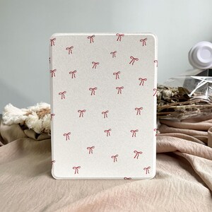 May include: An off-white tablet case featuring a repeating pattern of small, red bows. The case has a smooth surface and rounded corners. The background is a soft, neutral colour.
