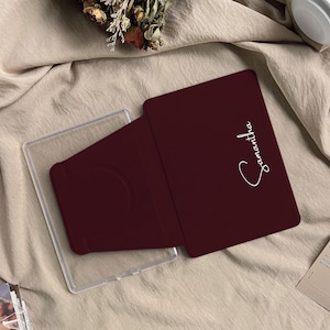 May include: A burgundy tablet case with the name "Samantha" in white script. The case is open, revealing a clear plastic holder. The case is on a beige fabric surface.