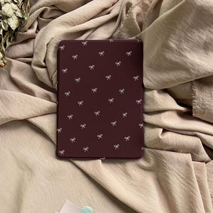 Puede incluir: Una funda rectangular para lector electr&oacute;nico en color burdeos intenso con un patr&oacute;n repetitivo de peque&ntilde;os lazos blancos dibujados con l&iacute;neas finas, distribuidos por toda la superficie. La funda tiene un acabado mate y liso.