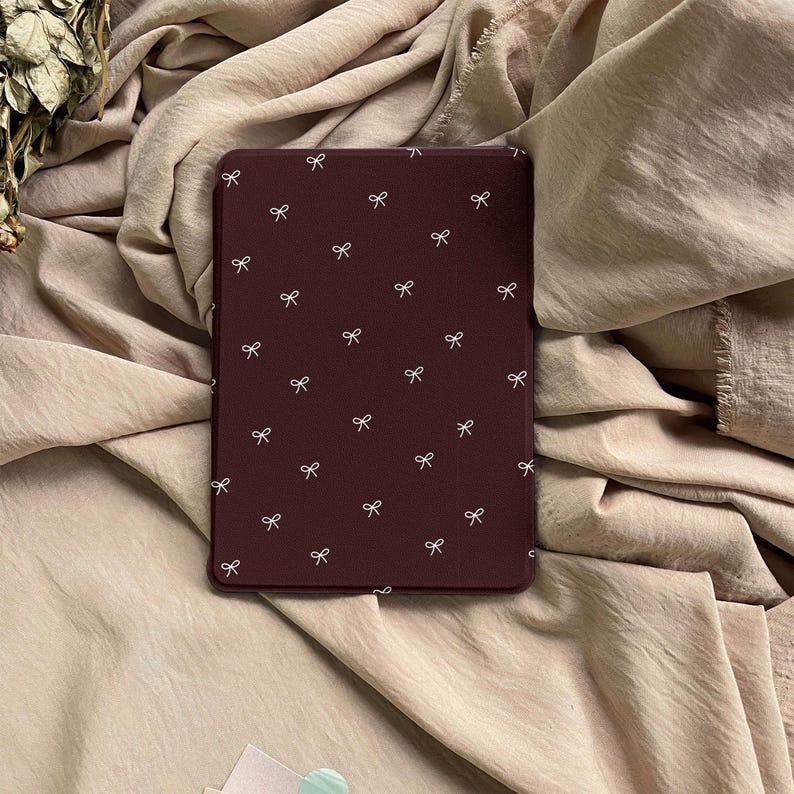 Puede incluir: Una funda rectangular para lector electr&oacute;nico en color burdeos intenso con un patr&oacute;n repetitivo de peque&ntilde;os lazos blancos dibujados con l&iacute;neas finas, distribuidos por toda la superficie. La funda tiene un acabado mate y liso.