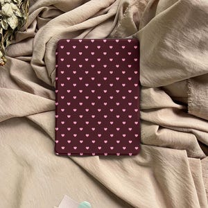 May include: A rectangular e-reader case featuring a dark maroon background with a repeating pattern of small, light pink hearts arranged in a grid. The case has rounded corners and a smooth, flat surface, displaying a consistent heart design.