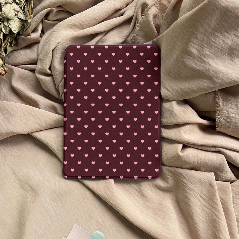 May include: A rectangular e-reader case featuring a dark maroon background with a repeating pattern of small, light pink hearts arranged in a grid. The case has rounded corners and a smooth, flat surface, displaying a consistent heart design.