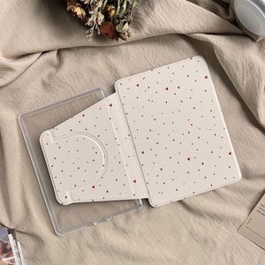 Puede incluir: Una funda de tableta de color crema con un patrón de pequeños corazones rojos. La funda está abierta, revelando un tarjetero de plástico transparente y el interior de la cubierta de la tableta. El fondo es una tela beige neutra.