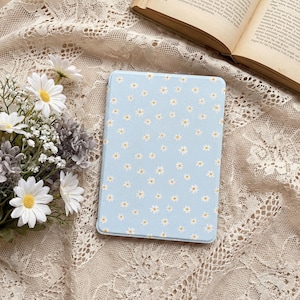 Puede incluir: Una funda rectangular para lector electrónico con fondo azul claro y un patrón repetitivo de pequeñas margaritas blancas con centros amarillos. La funda tiene una superficie lisa y plana con esquinas redondeadas para proteger el dispositivo.
