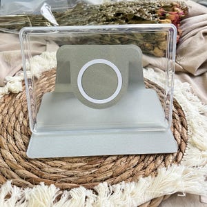 May include: A clear rectangular acrylic frame containing a light grey magnetic charging stand. The stand features a circular white ring centred on a raised, angled support platform, designed to hold a mobile device in place.