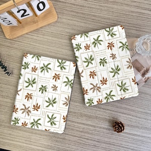 May include: Two white tablet cases with a repeating pattern of green and brown palm trees within a square grid. The cases are on a light wood surface, with a wooden calendar and a pinecone nearby.