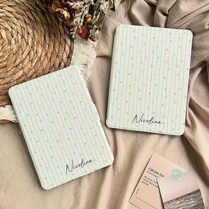 Puede incluir: Dos fundas rectangulares para lector electrónico con fondo crema claro, rayas verticales azul claro y pequeñas estrellas multicolores. Cada funda está personalizada con el nombre Nicolina en una fuente cursiva negra en el centro inferior.
