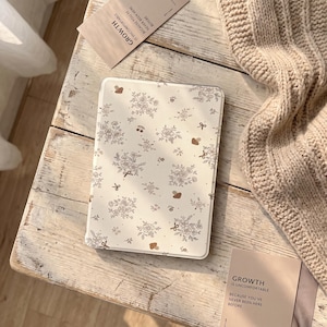 Könnte beinhalten: Eine rechteckige Tablethülle mit cremefarbenem Hintergrund und einem wiederkehrenden Muster aus braunen, liniengezeichneten Blumensträußen, kleinen Schleifen und Pilzen. Die Hülle liegt auf einer verwitterten weißen Holzoberfläche neben einer beigen Strickdecke.