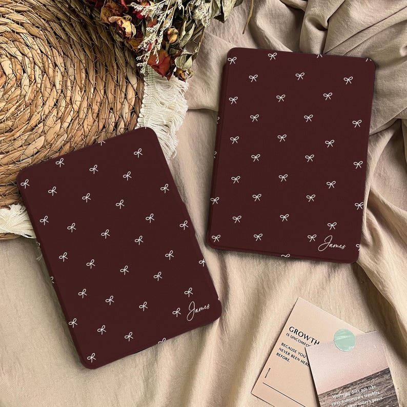 Puede incluir: Dos fundas rectangulares para lector electr&oacute;nico de color granate oscuro, con un patr&oacute;n repetitivo de peque&ntilde;os lazos blancos. Cada funda est&aacute; personalizada con el nombre James escrito en letra cursiva blanca en la esquina inferior derecha.