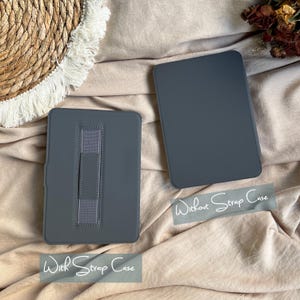 May include: Two dark grey rectangular e-reader cases. One case features a vertical elastic hand strap on the back, labelled With Strap Case. The other case has a smooth, plain back, labelled Without Strap Case. Both are solid-coloured.