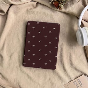 Puede incluir: Una funda para lector electr&oacute;nico de color granate oscuro con un patr&oacute;n repetitivo de peque&ntilde;os lazos blancos. La funda es rectangular con esquinas redondeadas y est&aacute; junto a auriculares blancos y una tarjeta con el texto GROWTH IS UNCOMFORTABLE.