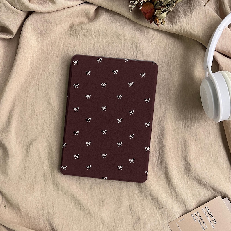 Puede incluir: Una funda para lector electr&oacute;nico de color granate oscuro con un patr&oacute;n repetitivo de peque&ntilde;os lazos blancos. La funda es rectangular con esquinas redondeadas y est&aacute; junto a auriculares blancos y una tarjeta con el texto GROWTH IS UNCOMFORTABLE.