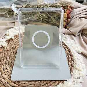 May include: A clear, rectangular plastic phone stand featuring a sage green interior panel with a white circular magnetic ring centred on the back support. The stand has a flat base and an upright frame designed to hold a mobile device securely.
