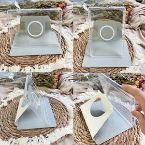 Puede incluir: Un soporte para tel&eacute;fono de acr&iacute;lico transparente con una base de carga magn&eacute;tica gris claro y un dise&ntilde;o de anillo blanco. El soporte cuenta con una base plegable de color gris claro y un marco transparente para dispositivos.