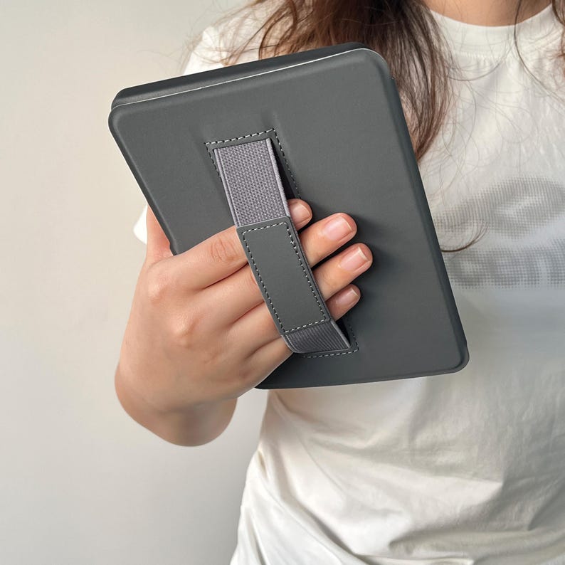 May include: A dark grey e-reader protective case featuring a built-in elastic hand strap on the back. The case has a smooth, matte finish with reinforced stitching along the edges of the strap, designed to provide a secure grip for the device.