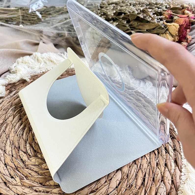 May include: A clear plastic phone case featuring a circular magnetic ring on the back and a foldable, cream-coloured stand attached to the interior. The stand has a large circular cutout and is designed to prop the case up at an angle.