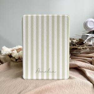 Puede incluir: Una funda rectangular con un patrón de rayas verticales en verde claro y blanco. El nombre "Amelia" está escrito en cursiva en la parte inferior. La funda es probablemente para una tableta o un lector de libros electrónicos.