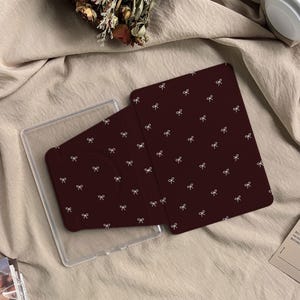 Puede incluir: Una funda para tableta de color granate oscuro con un patr&oacute;n repetitivo de peque&ntilde;os lazos blancos. La funda est&aacute; parcialmente abierta, revelando una carcasa de pl&aacute;stico transparente dise&ntilde;ada para sujetar el dispositivo.