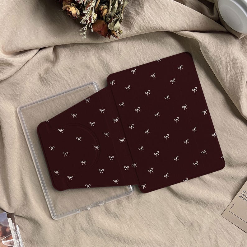 Puede incluir: Una funda para tableta de color granate oscuro con un patr&oacute;n repetitivo de peque&ntilde;os lazos blancos. La funda est&aacute; parcialmente abierta, revelando una carcasa de pl&aacute;stico transparente dise&ntilde;ada para sujetar el dispositivo.