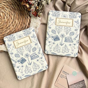 Op de afbeelding: Twee witte notitieboekjes met blauwe bloemmotieven en de naam "Jennifer" in gouden letters. De notitieboekjes bevatten illustraties van bloemen, aardbeien, typemachines en boeken. De notitieboekjes liggen op een beige ondergrond.