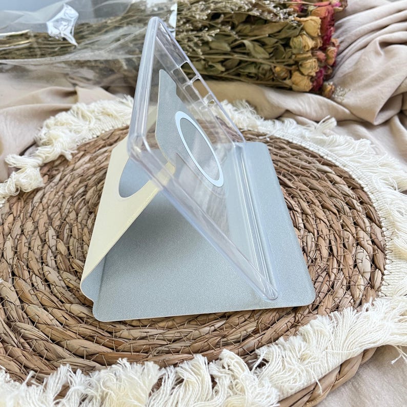 May include: A clear, foldable phone stand with a circular magnetic ring on the back panel. The stand features a light beige exterior and a textured grey interior base, designed to prop up a mobile device at an angled position.