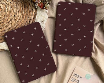 May include: Two rectangular e-reader cases in a deep maroon colour, each featuring a repeating pattern of small white line-drawn bows. The cases are positioned on a textured fabric surface, with one case placed vertically and the other slightly angled.