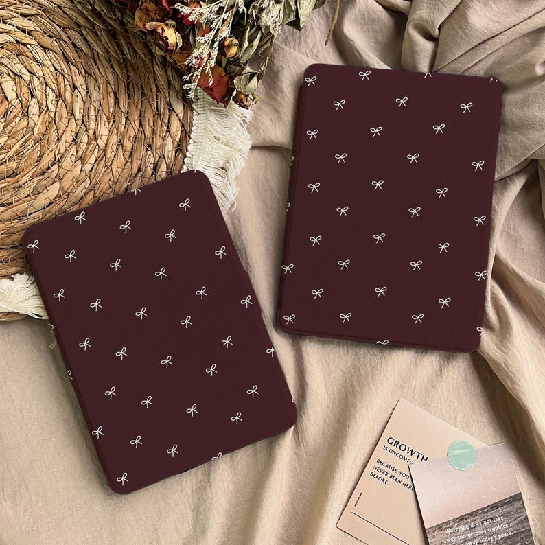 Puede incluir: Dos fundas rectangulares para lector electr&oacute;nico de color granate oscuro, cada una con un patr&oacute;n repetitivo de peque&ntilde;os lazos dibujados en l&iacute;neas blancas. Las fundas est&aacute;n colocadas sobre una superficie de tela texturizada, una vertical y otra inclinada.
