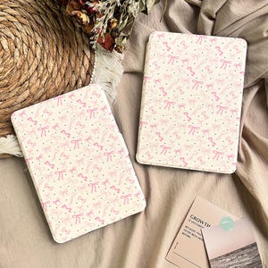 May include: Two white tablet cases with a repeating pattern of pink bows and small black dots. The cases are rectangular with rounded corners. The background is a neutral beige color.