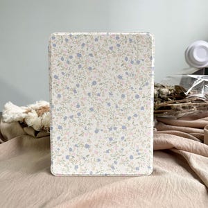 Puede incluir: Una funda rectangular para tableta con un estampado floral. La funda presenta un fondo color crema con un delicado patrón de pequeñas flores y enredaderas azules, rosas y verdes. La funda está diseñada para proteger una tableta y añade un toque de estilo.