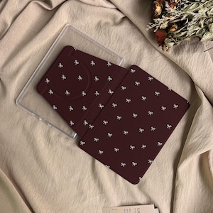 Puede incluir: Una funda para lector electr&oacute;nico o tableta de color granate oscuro con un patr&oacute;n repetitivo de peque&ntilde;os lazos blancos. La funda incluye un marco de pl&aacute;stico transparente y una cubierta granate con una abertura para la c&aacute;mara.