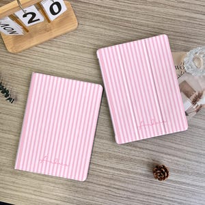 Może przedstawiać: Dwa różowo-białe etui na tablety w paski z nadrukiem "Andrea". Leżą na jasnej drewnianej powierzchni, z małą szyszką i drewnianym kalendarzem w tle.