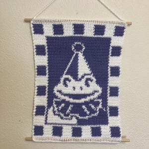 May include: Crocheted wall hanging featuring a frog wearing a party hat. The frog is white against a blue background, framed by a white and blue checkered border. The banner is suspended by a white string and wooden dowels.