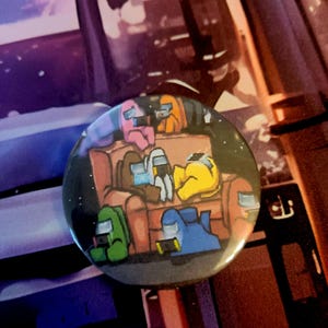 May include: A round button featuring a cartoon illustration of characters from the game Among Us. The characters, in various colors, are depicted relaxing on a brown couch. The background is a starry night.