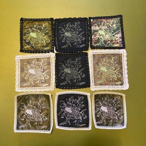May include: Nine square fabric coasters with floral embroidery. The coasters feature a variety of colours, including black, olive green, and tan, with white and gold embroidery. Each coaster has a decorative lace trim.